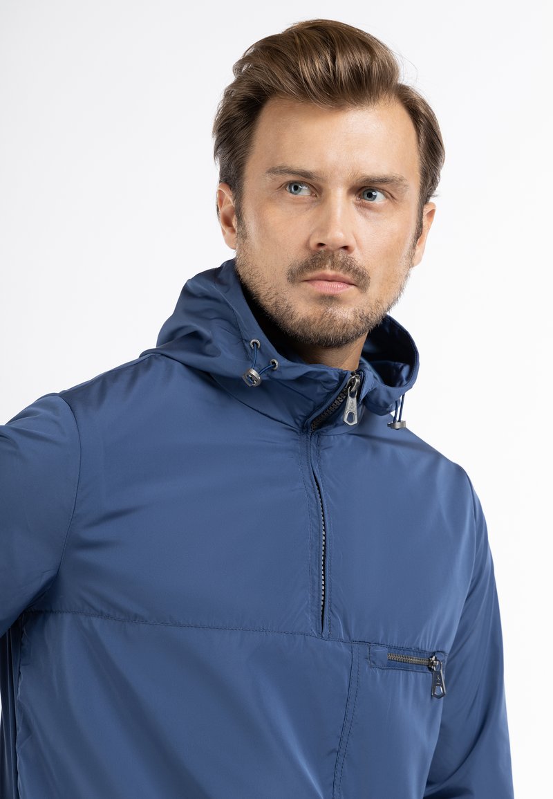 Veste bleu marine avec capuche, dotée d'un zip half-zip à l'avant, d'une petite poche zippée sur la poitrine et d'accents avec cordon de serrage sur la capuche. Texture lisse.