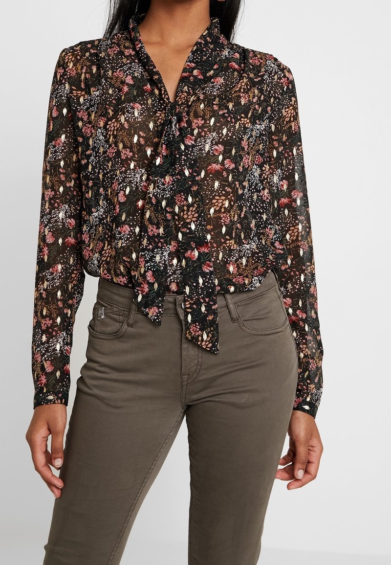 Blouse noire à motif floral avec un col noué, présentant des motifs de fleurs roses et blanches, associée à un pantalon vert olive, le tissu semble léger et texturé.