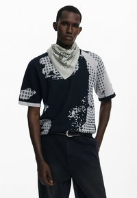 Kortærmet sweater med et sort og gråt abstrakt mønster med gitterelementer; parret med et lysmønstret bandana og mørke bukser.