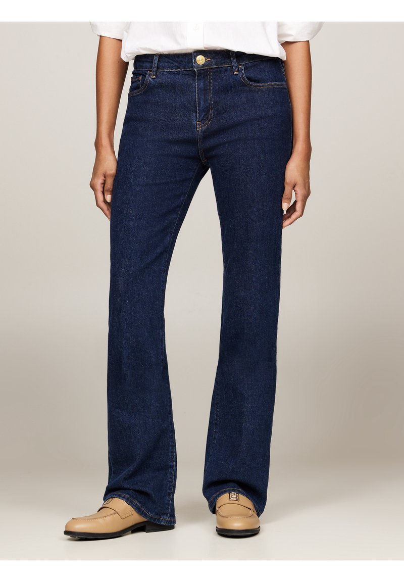 Persona con jeans rectos de color azul oscuro y mocasines beige, de pie contra un fondo claro y liso.