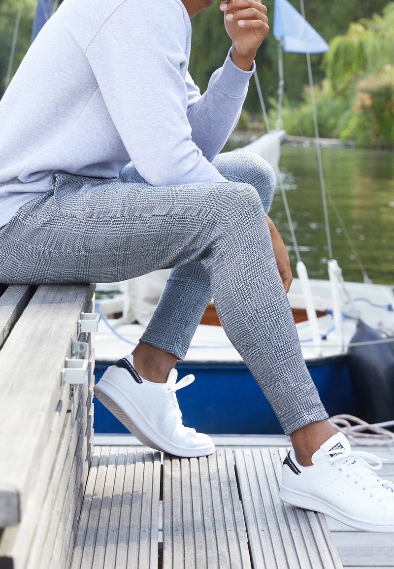 Szare leginsy w kratkę, wykonane z lekkiego materiału, zestawione z białymi sneakersami. Tło przedstawia pomost obok spokojnej tafli wody.
