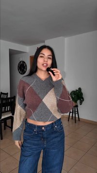 Jeune femme en pull à motifs et jean tient un pinceau de maquillage près de sa bouche dans un salon avec une plante et une horloge murale.