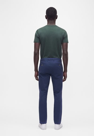 Homme debout de dos, portant une chemise verte à manches courtes, un pantalon bleu et des baskets blanches sur un fond clair uni.