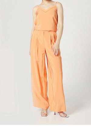 Kobieta w brzoskwiniowych spodniach o szerokich nogawkach i dopasowanym topie na cienkich ramiączkach, w towarzystwie białych sandałów na wysokim obcasie z paskami.