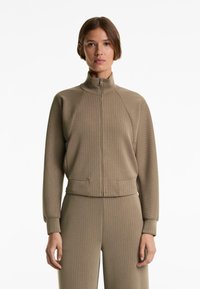 Giacca beige con zip, tessuto a coste, colletto alto e tasche laterali, abbinata a pantaloni a coste coordinati.
