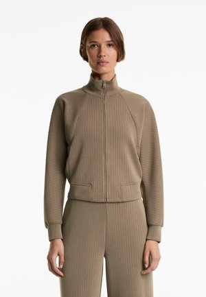 Beige ritsjack met ribbelstructuur, hoge kraag en zijzakken, gecombineerd met bijpassende ribbelbroek.