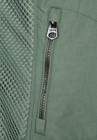 Světlounce zelená bunda z tkaniny s mesh panely, černým zipem a detaily šití. Povrch je hladký s mírně strukturovaným finišem.