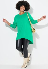 Grünes Langarm-Tunika mit einem High-Low-Saum, kombiniert mit schwarzen Leggings und glänzenden goldenen Cowboy-Stiefeln. Eine goldene Umhängetasche setzt einen Akzent.