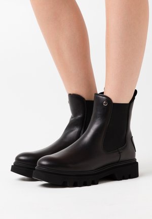 Černé kožené kotníkové boty s robustní podešví ve tvaru šroubů, nošené na bosých nohou, s elastickými bočními panely a malým kovovým knoflíkem jako detailem.