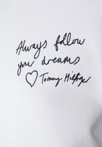 Sweatshirt en coton blanc avec du texte manuscrit noir : "Suivez toujours vos rêves" avec un cœur et la signature "Tommy Hilfiger".