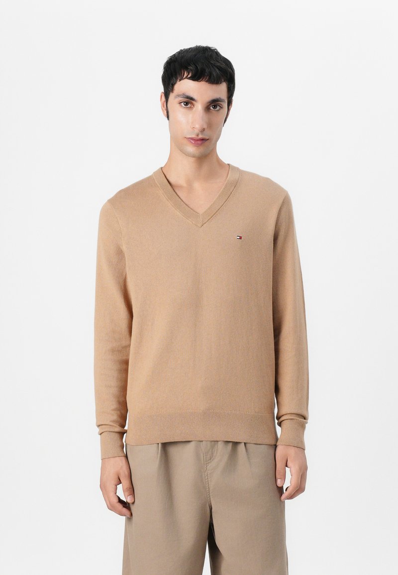 Beige V-hals trui gemaakt van zacht materiaal, met lange mouwen en een geribbelde onderkant. Kleine logodetail op de borst.