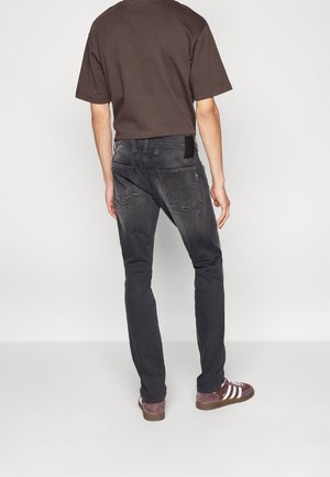 Mann trägt dunkel gewaschene Slim-Fit-Jeans, braunes T-Shirt und kastanienbraune Sneakers mit weißen Streifen, steht mit dem Rücken zur Kamera auf einem grauen Boden.