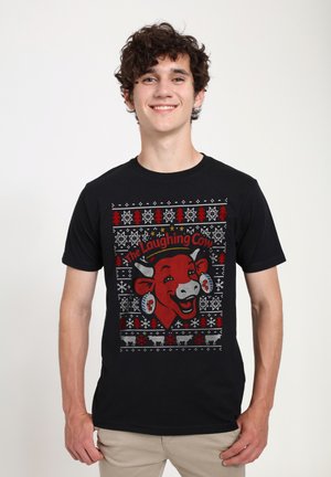 Sort T-shirt med et farverigt grafisk motiv af en smilende rød ko med accenter, omgivet af festlige snefnug og træer i et strikket mønster.
