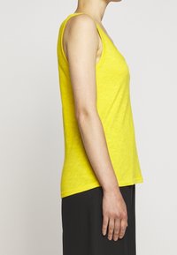 Keltainen hihaton paita, jossa on V-pääntie, valmistettu pehmeästä, teksturoituneesta kankaasta, löysä istuvuus ja suora helma, yhdistetty tummaan alaosaan.