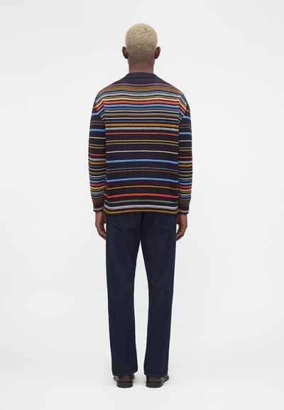 Άνδρας με κοντά ξανθά μαλλιά στέκεται με την πλάτη, φοράει πολύχρωμο πουλόβερ με οριζόντιες ρίγες, σκούρο παντελόνι και μαύρα παπούτσια.