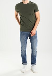 Maglietta a maniche corte verde oliva con colletto rotondo, abbinata a jeans in denim azzurro chiaro e sneakers bianche. Design semplice con un dettaglio logo.