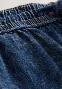 Material denim cu talie elastică strânsă. Culoare albastru închis cu textură subtilă și cusături aurii contrastante de-a lungul cusăturii.