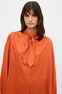 Orange-Bluse mit einem Bindeband-Ausschnitt, langen Ärmeln und Knopfverschluss. Sie hat eine glatte Textur und eine leicht lockere Passform.