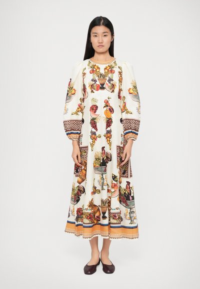 Femme portant une longue robe blanche à manches bouffantes, ornée de motifs détaillés de fruits colorés et de fleurs, associée à des chaussures plates foncées.