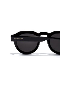Schwarze Sonnenbrille mit einem dicken, eckigen Rahmen. Dunkle Gläser und das Logo "HAWKERS" in Gold an der Seite. Glatte, glänzende Oberfläche.