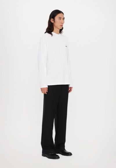 Cămașă albă cu mânecă lungă, croi lejer, cu logo din text negru. Potrivita cu pantaloni negri, plisati, cu picioare largi și pantofi negri. Design simplu și curat.