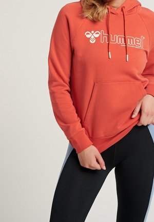 Frau trägt einen orangen Hummel-Hoodie mit Kängurutasche und schwarze Leggings mit hellblauen Seitenstreifen, steht vor einem neutralen Hintergrund.