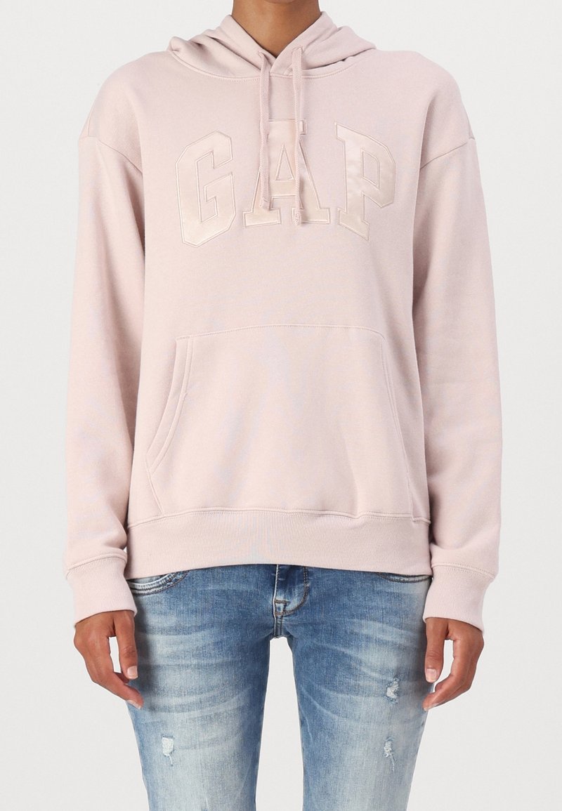 Sweat à capuche rose clair avec poche avant et logo « GAP » associé à un jean bleu délavé déchiré sur une personne debout.
