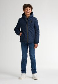 Dunkelblaues Puffer-Jacke mit Kapuze, Reißverschluss, elastischen Bündchen und einem Logo-Patch auf der Brust, kombiniert mit blauen Jeans und weißen Sneakers.