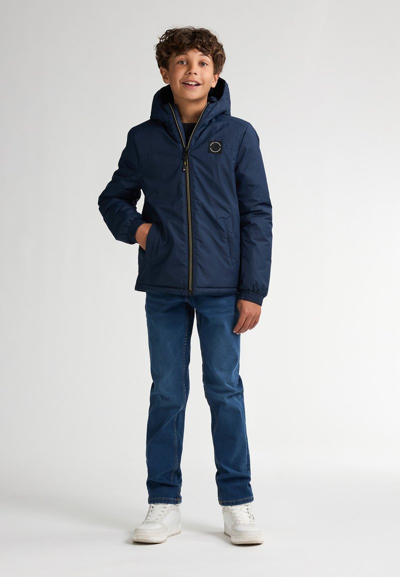 Dunkelblaues Puffer-Jacke mit Kapuze, Reißverschluss, elastischen Bündchen und einem Logo-Patch auf der Brust, kombiniert mit blauen Jeans und weißen Sneakers.