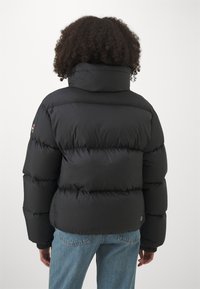 Fekete puffertöltetű dzseki magas gallérral és steppelt mintával. Rugalmas mandzsettákkal és egy kis logós részlettel a hátsó részen. Kék farmernadrág felett viselve.