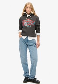 Sudadera corta gris oscuro con cordones blancos y un gráfico floral. Llevada sobre una camisa blanca, combinada con jeans azul claro y zapatos negros.