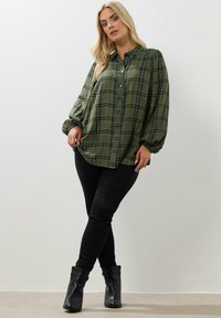 Zielona koszula w kratę z guzikiem i marszczonymi rękawami, noszona na czarnych skinny jeansach i czarnych botkach za kostkę. Miękka tkanina, luźny krój, styl casual.