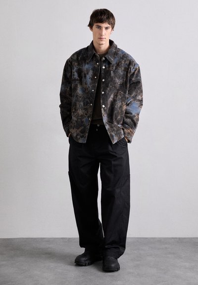 Button-up jacket with a dark, patterned fabric featuring blue and brown hues; paired with loose-fitting black cargo pants and black shoes.