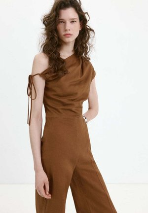 Junge Frau mit lockigem Haar trägt einen braunen One-Shoulder-Jumpsuit mit gebundenem Träger und weitem Bein, steht vor weißem Hintergrund.