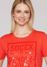 Czerwona bawełniana koszulka z krótkim rękawem i okrągłym dekoltem, z błyszczącym czarnym nadrukiem na przodzie, zawierającym napis "SOCCX a RENDEZ-VOUS" umieszczony na środku.