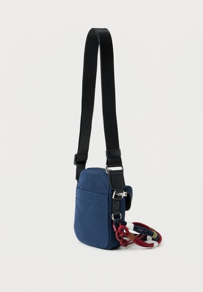 Μικρό τσαντάκι χιαστί σε μπλε ναυτικό χρώμα με μαύρο ρυθμιζόμενο λουράκι και πολύχρωμο πλεκτό μπρελόκ στο πλάι, σε λευκό φόντο.