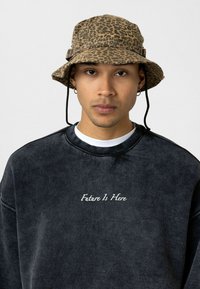Bruine panterprint emmerhoed met verstelbare kinriem; zachte zwarte sweatshirt met witte tekst "Future Is Here" op de voorkant.
