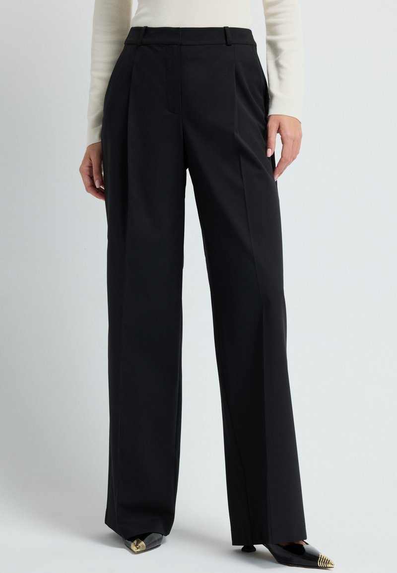 Zwarte wijd uitlopende broek met hoge taille en plooien, gemaakt van een soepele stof, gecombineerd met zwarte schoenen met spitse neuzen en gouden accenten.