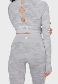 Hellgraues, kurz geschnittenes Langarmoberteil mit kreisförmigen Ausschnitten und passenden hochgeschnittenen Leggings, beide im dezenten Camouflage-Muster.