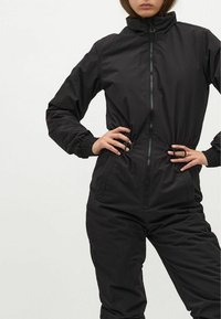 Schwarzer Overall mit hohem Kragen, Frontreißverschluss, elastischen Manschetten und Kordelzugbund. Hergestellt aus glattem, leichtem Stoff.