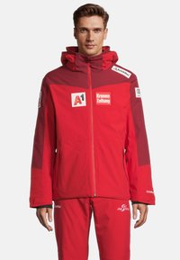 Rote Skijacke mit Kapuze, Reißverschluss und mehreren Sponsorenlogos. Das Material scheint wasserabweisend zu sein; das Design umfasst kontrastierende Einsätze.