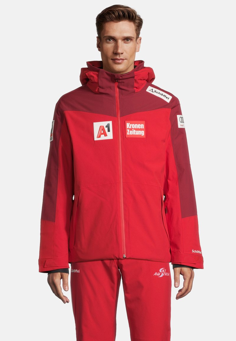 Rote Skijacke mit Kapuze, Reißverschluss und mehreren Sponsorenlogos. Das Material scheint wasserabweisend zu sein; das Design umfasst kontrastierende Einsätze.