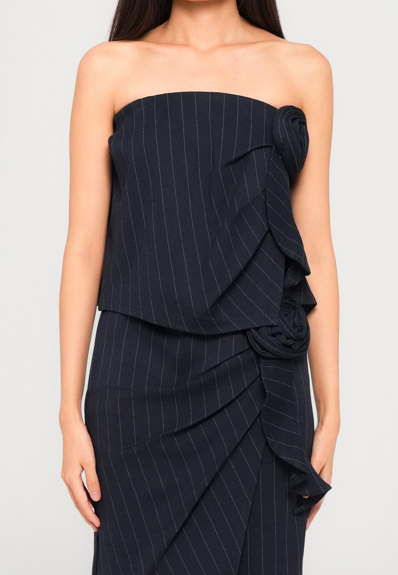 Femme portant une robe bustier bleu marine à fines rayures avec des volants superposés et des détails de roses en tissu sur la poitrine et la hanche.