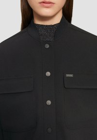 Czarna kurtka z gładkiego materiału, z stójką, dwoma kieszeniami na piersi, zatrzaskami i subtelnym akcentem logo.