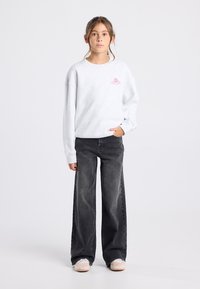 Tyttö seisoo, yllä valkoinen huppari, jossa on vaaleanpunainen logo, mustat väljät farkut ja vaaleanpunaiset skeittikengät yksinkertaisella vaalealla taustalla.