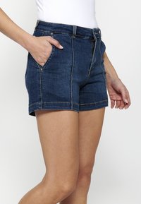 Denim kratke hlače v temno modri barvi s prilegajočim dizajnom, ki vključujejo dva sprednja žepa, kontrastno šivanje ter zadrgo in gumb za zapiranje.