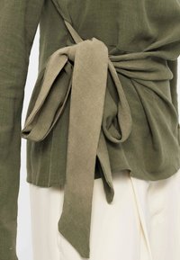 Haut à manches longues vert olive avec une large ceinture en tissu nouée en nœud à la taille sur un pantalon écru.