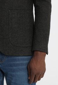 Tmavě šedý sako ve vzoru herringbone s texturovanou tkaninou, které má jednu náprsní kapsu, klopové límce a mírně zúžené rukávy.