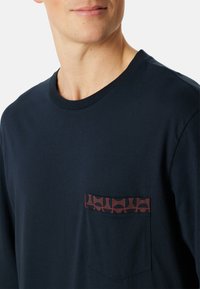 Marinblå bomulls t-shirt med rund halsringning och en bröstficka som har ett geometriskt mönster i rött och krämfärgat. Slät textur.