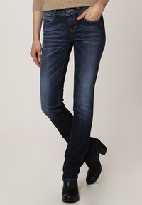 Jeans de mezclilla azul oscuro con corte recto, cinco bolsillos y detalles de costura visibles. Combinados con botas de tobillo negras.